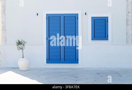 Blaue Fensterläden schmücken eine weiße mediterrane Hausfassade mit Olea europaea in einem Topf. Konzept von Architektur, Harmonie und Küstenleben Stockfoto