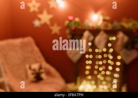 Unscharfer Schuss. Verschwommener Weihnachts-Hintergrund mit Sessel und Kamin, dekoriert mit hängenden Socken, Tannenzweigen, roten Kugeln und goldener Girlande Stockfoto