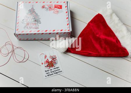 Ismail, Ukraine – 13. Dezember 2019. Weihnachtsgeschenk, Weihnachtsmannmütze auf weißem Holztisch. Aufschrift auf der Karte: Glück ist Schnee, Süßigkeiten und warme Handschuhe Stockfoto