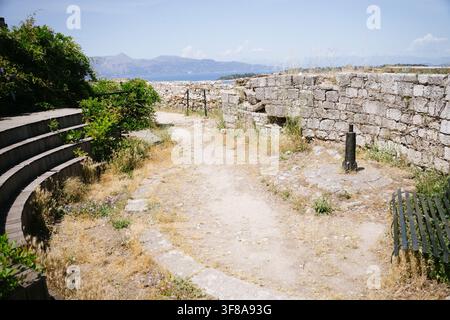 Ein Feldweg windet sich zwischen einer alten Steinmauer der Festung und hölzernen Amphitheaterstufen mit Blick auf das Meer und die Berge in Korfu, Griechenland Stockfoto