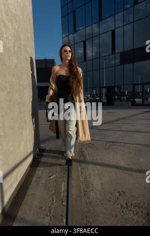 Stylische junge Frau geht selbstbewusst in der Nähe eines modernen Glasgebäudes. Modisches Model trägt beigen Trenchcoat, Sonnenbrille und Jeans im Freien. Trend Stockfoto