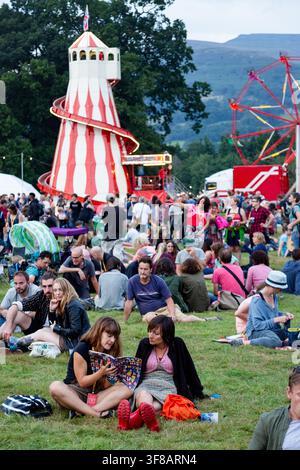 FREUNDE LESEN DAS PROGRAMM AUF DEM GREEN MAN FESTIVAL, REPORTAGE, GREEN MAN FESTIVAL 2016, BRECON, WALES, UK: Zwei junge Frauen sitzen auf dem Gras und teilen ein Buch, während ein farbenfroher Rutsche und ein Riesenrad hinter ihnen auf einem Festival sichtbar sind. Foto: Rob Watkins. INFO: Das Green Man Festival begann 2003 in Wales. Jährlich findet es im dritten Augustwochenende in den Brecon Beacons statt und feiert alternative Musik, Kunst, Literatur, Wissenschaft und familienfreundliche Kultur in einer wunderschönen Berglandschaft. Stockfoto
