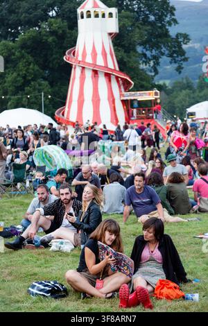 FREUNDE LESEN DAS PROGRAMM AUF DEM GREEN MAN FESTIVAL, REPORTAGE, GREEN MAN FESTIVAL 2016, BRECON, WALES, UK: Zwei junge Frauen sitzen auf dem Gras und teilen ein Buch, während ein farbenfroher Rutsche und ein Riesenrad hinter ihnen auf einem Festival sichtbar sind. Foto: Rob Watkins. INFO: Das Green Man Festival begann 2003 in Wales. Jährlich findet es im dritten Augustwochenende in den Brecon Beacons statt und feiert alternative Musik, Kunst, Literatur, Wissenschaft und familienfreundliche Kultur in einer wunderschönen Berglandschaft. Stockfoto
