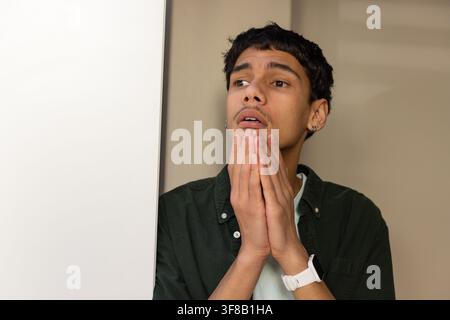 Erwachener afroamerikanischer Mann, der die Hände nahe am Kinn an einer weißen Tafel drückt, trägt ein grünes Hemd und eine Uhr. Porträt, Solo, nachdenklich, minimal, beige, Ohr Stockfoto