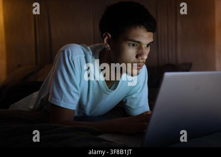 Afroamerikaner liegt auf dem Bett, trägt ein weißes T-Shirt und benutzt einen Laptop im Schlafzimmer mit Lampenschein. Dämmerig, gemütlich, intim, stimmungsvoll, abgelegen, Arbeit, Konzentration Stockfoto