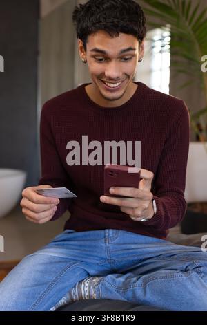 Afroamerikanischer Mann in den 20ern trägt einen burgunderroten Pullover, hält ein Smartphone und eine Karte auf einem Kissen zu Hause. Fintech, Zahlung, Mobil, Transaktion, Geldbörse, App Stockfoto