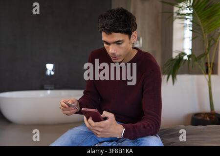 Afroamerikaner sitzt auf einer gepolsterten Bank in einem modernen Badezimmer und hält ein Smartphone und eine Bankkarte. Gentleman, Telefon, Zahlung, Brieftasche, Pflanze, natürlich Stockfoto