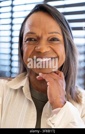 Afrikanische Seniorin lächelt, ruht das Kinn, trägt ein Hemd mit Kragen, olivgrünes Oberteil, Armband, zu Hause vor Jalousien. Porträt, betagt, Wärme, natürlich, Licht, Fenster Stockfoto