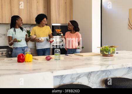 Drei afrikanische Frauen stehen hinter einer Marmorinsel in der Küche und bereiten einen Salat mit Paprika zu, während sie Jeans tragen. Teamarbeit, Kochen, Freundschaft, Gemüse, Tageslicht Stockfoto