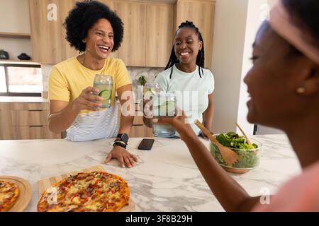 Afrikanische Frauen, die Gläser mit Gurkenwasser halten und an einer Marmorinsel mit Pizzen und Salat anstoßen. Modern, Küche, sozial, Freunde, Zusammenkunft, Theke Stockfoto