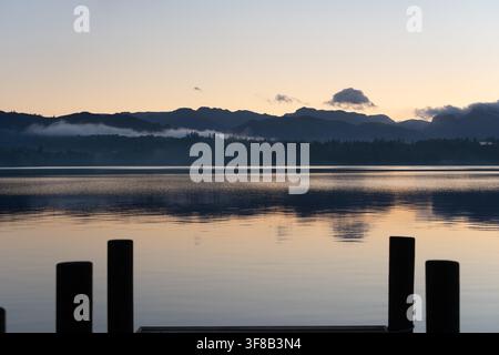 Nebel legt sich über Windermere im Lake District in der Morgendämmerung, gesehen von einem Steg mit den ikonischen Langdale Pikes in der Ferne. Stockfoto
