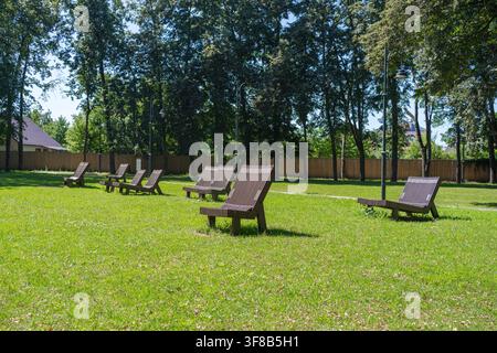 Öffentlicher Ruheplatz in einem Stadtpark mit ergonomischen Holzliegestühlen auf grünem Gras, umgeben von Bäumen. Ideal für den Sommerurlaub, urbaner Komfort Stockfoto