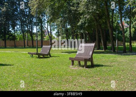 Holzliegestuhl steht auf frischem grünem Gras im Schatten von Bäumen in einer Stadtparkzone. Perfektes Bild für Outdoor-Lebensstil, umweltfreundliche Stadt Stockfoto