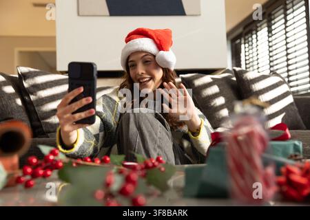 Frau sitzt auf der Couch zu Hause, trägt einen Weihnachtsmannhut und einen Strickpullover, hält ein Smartphone, winkt. Feiertag, festlich, Telefon, Geschenke, Tageslicht, gemütlich, modern Stockfoto