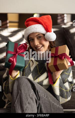 Frau sitzt auf dem Sofa und hält zwei verpackte Geschenke, trägt einen roten Weihnachtsmannhut im Wohnzimmer. Geschenke, festlich, gemütlich, Inneneinrichtung, Argyle, fröhlich, Lebensstil Stockfoto