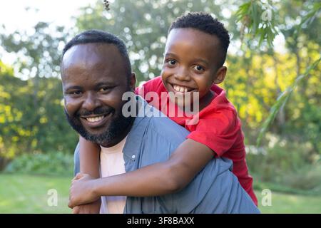 Afroamerikanischer Vater und Sohn lächeln, teilen ein Tragetuch im sonnendurchfluteten Graspark, hellblaues Hemd. Bärtig, Outdoor, Familie, Porträt, Bindung, Kandel Stockfoto