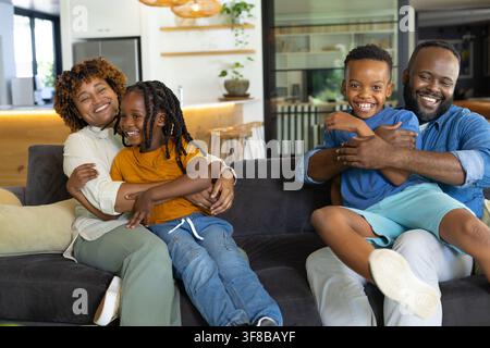 Afroamerikanische Familie sitzt auf grauem Sofa, umarmt Kinder mit Kücheninsel-Pendelleuchten. Zuhause, Einrichtung, Wärme, zusammen, gemütlich, modern, offene Planung Stockfoto