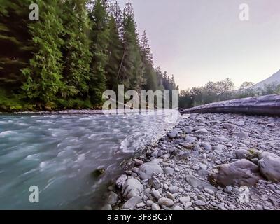Ein rauschender Fluss fließt über glatte Steine. Üppige immergrüne Bäume säumen das Flussufer. Der Himmel leuchtet sanft mit Dämmerungsfarben. Felsen glitzern mit frischem Flusssprühregen. N Stockfoto