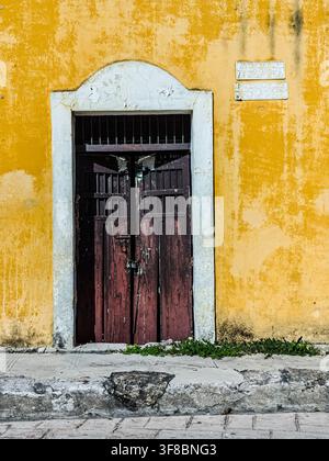 Ansicht einer verwitterten dunklen Holztür, die in eine leuchtend gelbe Stuckwand mit einem weißen gebogenen Rahmen in Izamal, Yucatan, Mexiko, eingelassen ist. Stockfoto