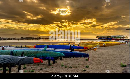 Goldener Sonnenaufgang über dem Strand mit Reihen von Kajaks im Sand, mit dem historischen Stearns Wharf und dramatischen Sonnenstrahlen, die durch die Wolken brechen. Stockfoto
