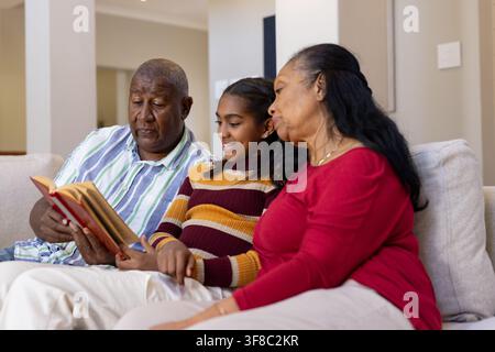 Afroamerikanische Familie liest rotes Buch auf hellem Sofa im Wohnzimmer, Mädchen in gestreiftem Pullover Stockfoto