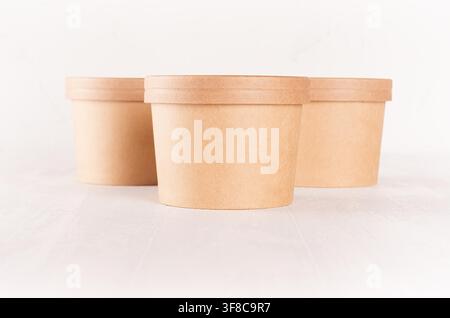 Gruppe von drei leeren braunen Einwegkartons für Essen zum Mitnehmen mit Kappen - Suppe, Salat, Eis auf weißem Holzregal in der Nähe. Stockfoto