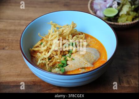 Traditionelle nordthailändische Kokosnuss-Kurry-Nudelsuppe mit Hühnerdrumstick, garniert mit knusprigen Eiernudeln in einer blauen Schüssel. Stockfoto