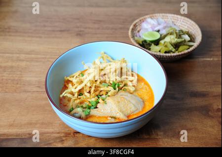 Traditionelle nordthailändische Kokosnuss-Kurry-Nudelsuppe mit Hühnerdrumstick, garniert mit knusprigen Eiernudeln in einer blauen Schüssel. Stockfoto