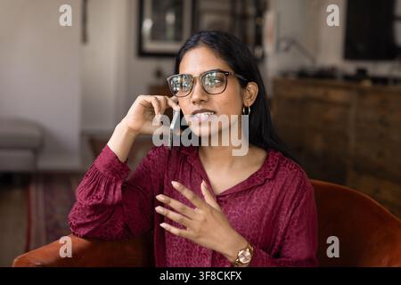 Nahaufnahme einer jungen indischen Frau mit Brille, die auf einem Sessel sitzt, ein Telefonat zu Hause führt, ein Ferngespräch mit einem Familienmitglied führt, über Persisches spricht Stockfoto