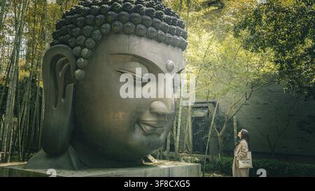 Frau blickt zu einem riesigen Buddha-Kopf-Denkmal in einem Bambuswald im Botanischen Garten von Hangzhou auf Stockfoto