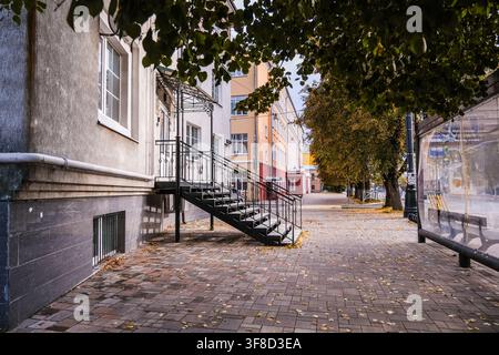 Rjasan, Russland, 28.09.2025 Leerer Gehweg mit gefallenen Herbstblättern und einer Außentreppe aus Metall, die zu einem Gebäudeingang führt, mit einer Bushaltestelle in der Nähe Stockfoto