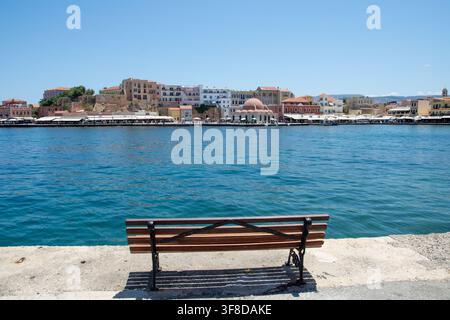 Blick auf die Altstadt-Marina von Chania mit einer hölzernen Bank im Vordergrund, historischen Gebäuden und der Kuppel der Kucuk-Hasan-Moschee über dem blauen Hafenwasser Stockfoto