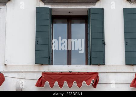 Ein hohes Glasfenster mit offenen grünen Fensterläden ist in einer weißen Fassade über einem roten Markisenvordach eingelassen. Stockfoto