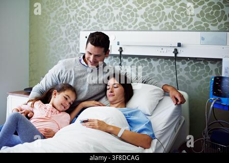 Lächeln, Familie und Frau im Bett im Krankenhaus, Unterstützung und Bindung für die medizinische Genesung. Vater, Kind und Besuch der Mutter auf der Intensivstation für Glück Stockfoto