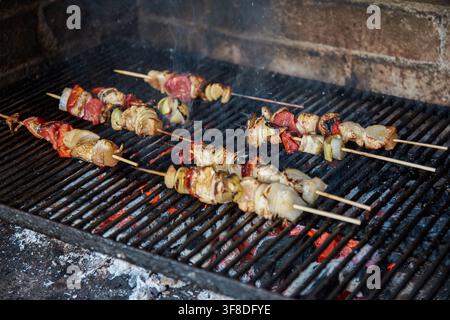 Leckere Shish-Kebap-Spieße mit marinierten Fleischstücken, Zwiebeln und Tomaten, die über heiß glühender Kohle auf einem Metallrost in einem Ziegel-Kamin gegrillt werden Stockfoto
