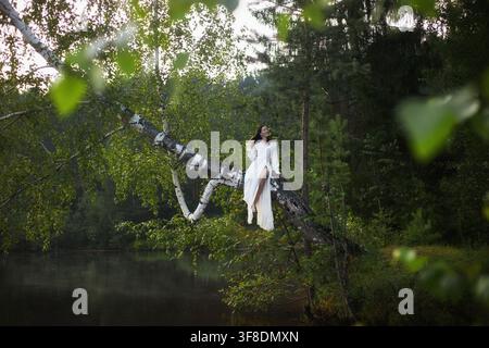 Frau in einem weißen Outfit sitzt auf einem großen Ast über einem ruhigen See in einem Wald. Die Sonne scheint durch die Bäume und wirft Schatten auf das Wasser. Stockfoto