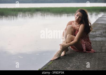 junge Frau sitzt auf einem steinernen Dock an einem Gewässer. Sie hat einen nachdenklichen Blick im Gesicht. Das Wasser ist ruhig, und der Himmel ist leicht bewölkt in der Nachmittagsstunde Stockfoto