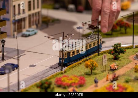 Eine Nahaufnahme zeigt eine erwachsene Hand, die eine Miniaturstraßenbahn auf einem detaillierten Stadtdiorama mit Gleisen, Autos, Gebäuden, Bäumen und Parkwegen einstellt. Stockfoto