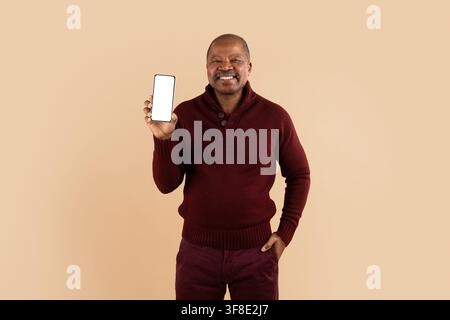 Lächelnder reifer schwarzer Mann hält ein leeres Smartphone, während er auf beigem Hintergrund steht Stockfoto