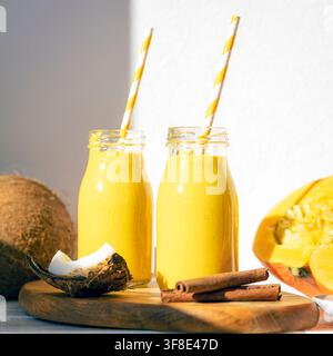 Kürbis- und Kokosnuss-Smoothies in Glasflaschen mit gestreiften Strohhalmen, frische Kokosnuss, Mango und Zimt auf einem Holzbrett, Nahaufnahme Stockfoto