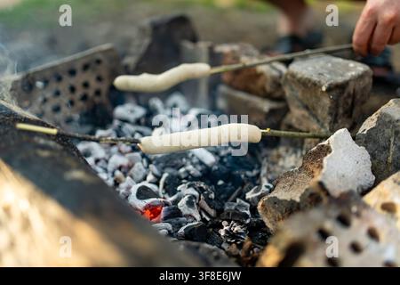 Würste braten an Holzspießen über heißen Lagerfeuer-Glut im Freien. Traditionelle Lagerfeuer-Kochkunst schafft eine einfache Mahlzeit beim Camping und zur Erholung Stockfoto