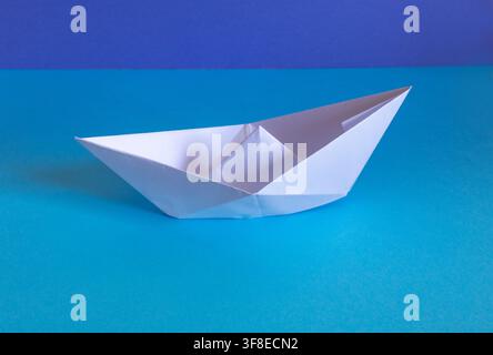 Einzelnes weißes Origami-Papierboot auf blauer Oberfläche mit tiefblauem Hintergrund Stockfoto