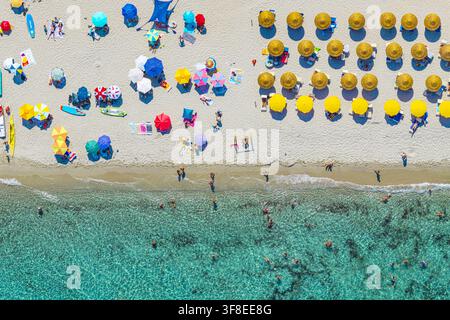 Luftbild von bunten Strandschirmen, die in ordentlichen Reihen auf dem weißen Sand angeordnet sind, mit Menschen, die im klaren türkisfarbenen Wasser des Mittelmeers schwimmen Stockfoto