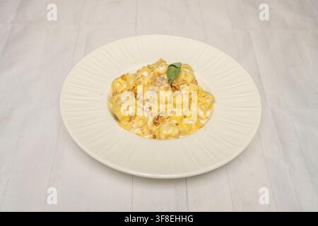 Kremiges Tortellini-Nudelgericht mit gehackten Walnüssen und einem frischen Salbeiblatt, serviert auf einem weißen gerippten Teller. Stockfoto