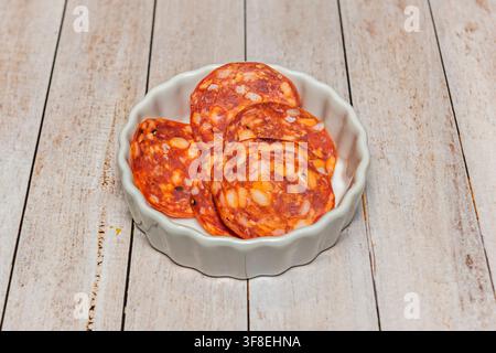 Geschnittene würzige Salami-Rondellen, die in einer kleinen geriffelten weißen Schale auf einem rustikalen Holztisch arrangiert sind. Stockfoto