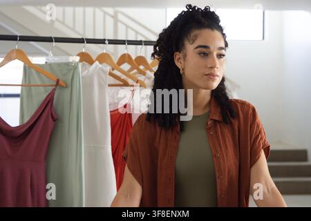 Female stylist steht rechts im Studio neben einem Kleiderständer mit Bügeln, trägt ein rostfarbenes Hemd und goldene Ohrringe Stockfoto