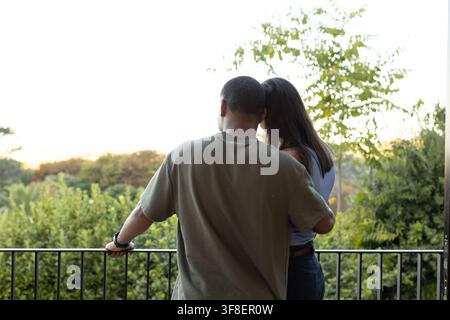 Afroamerikanisches Paar steht auf dem Balkon und beobachtet den Sonnenuntergang, Mann in olivgrünem T-Shirt zeigt Armbanduhr Stockfoto