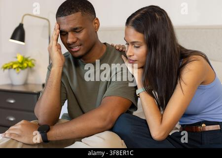 Afroamerikanisches Paar sitzt zu Hause auf dem Bett, Mann hält den Kopf, Frau berührt Smartwatch Stockfoto