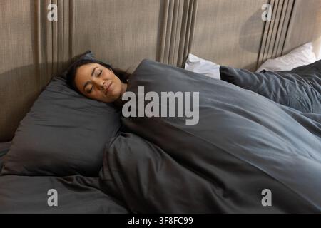 Erwachsene asiatische Frau liegt auf der Seite im Schlafzimmer unter einer dunkelgrauen Bettdecke mit Kissen Stockfoto