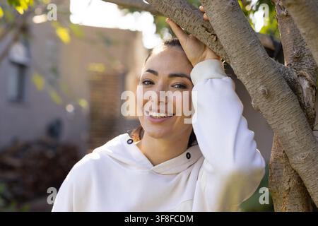 Frau lehnt sich an einen Baumstamm, ruht auf einem Ast, trägt einen hellen Hoodie in der Nähe von Stuckstufen Stockfoto